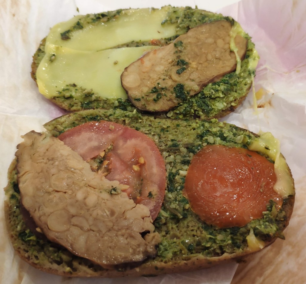 An opened sandwich with a brown bread bun covered in pesto with a couple of slices of cheese, two slices of tomato, and two slices of tempeh.