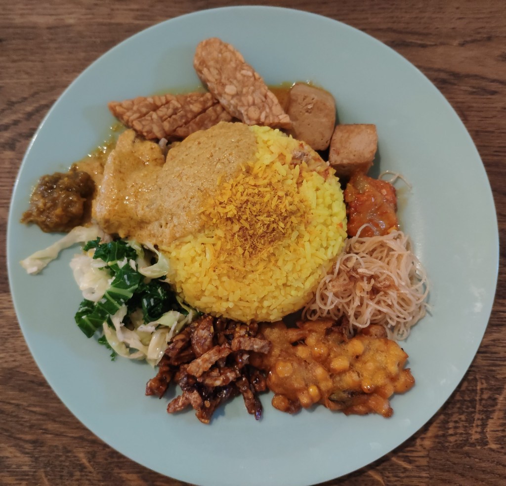 A mixed plate of Indonesian food. There is a big mound of rice in the middle of the plate, and lots of small dollops of different dishes around the edge.