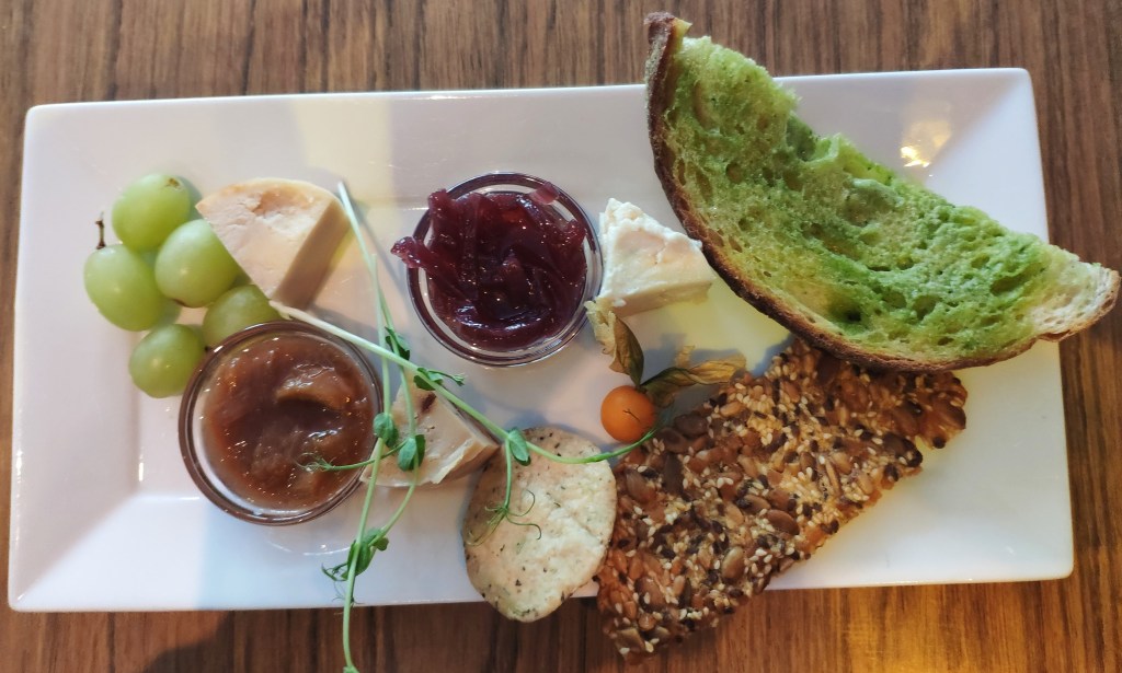 A cheese plate featuring one slice of bread, two crackers, three pieces of cheese, two chutneys, and some fruit,