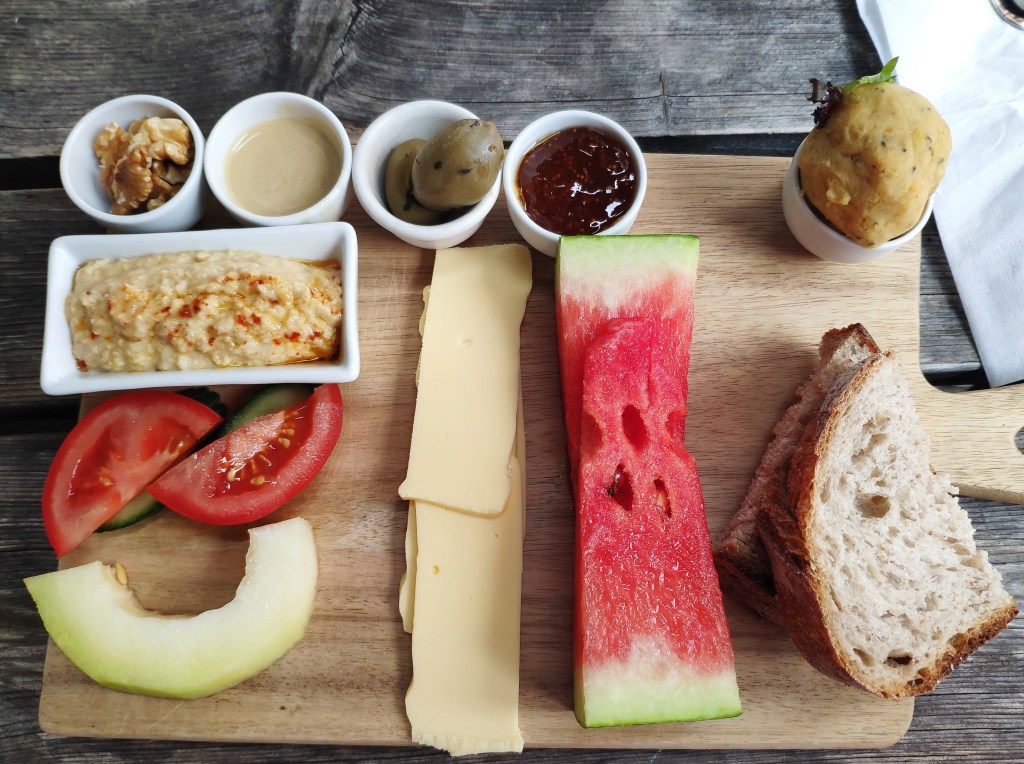 A board holding some sliced fruit and vegetables, a little bit of bread, and a few small pots containing different spreads.