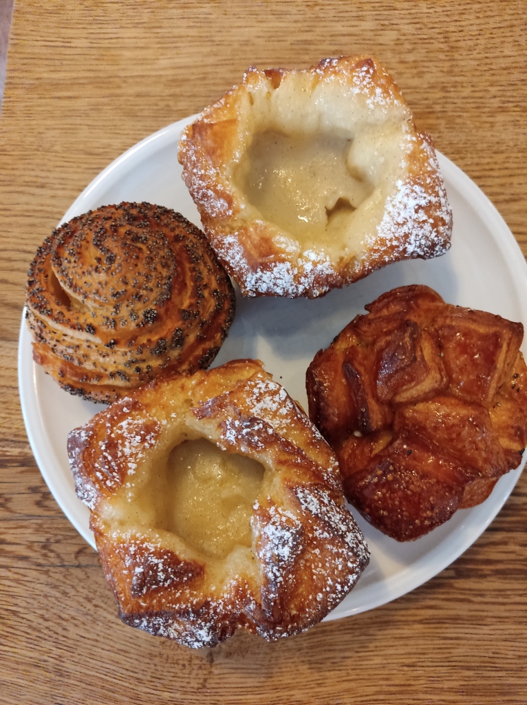 A plate of four pastries - two pastry cups filled with custard, a snail, and a bun with crusty pieces on top