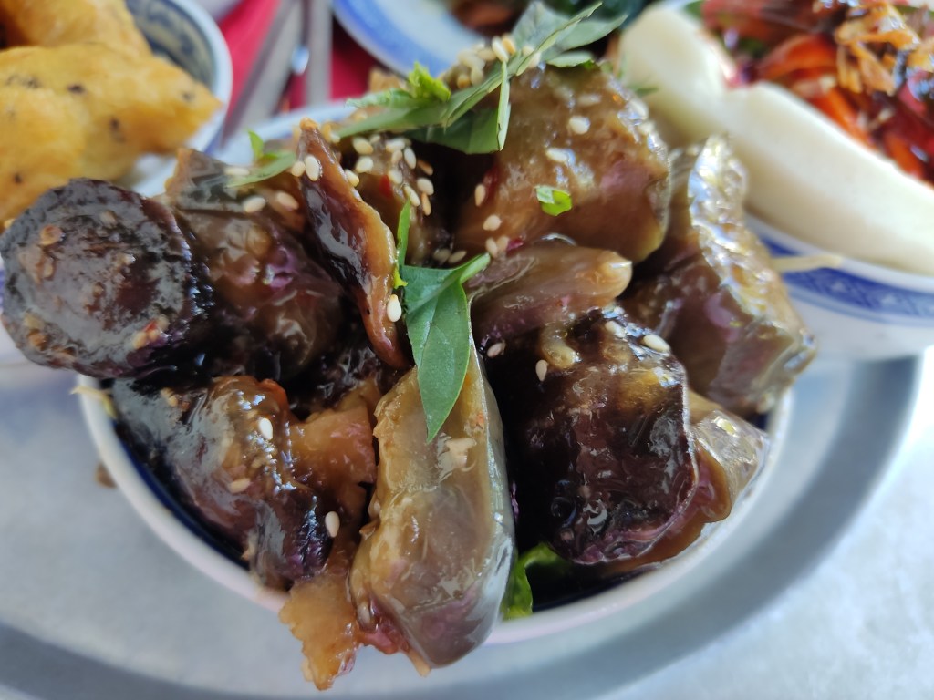 A small bowl overflowing with oily looking well cooked aubergine and mushroom, topped with sesame seeds and fresh herbs