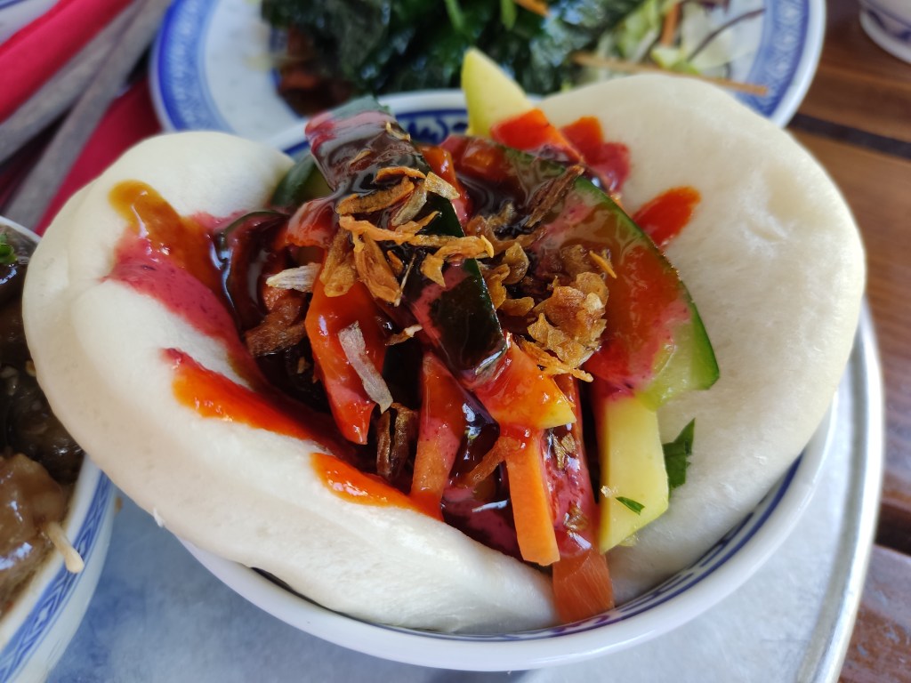 A bao bun filled with soy meat and stripes of fresh vegetables, topped with fried onion and drizzled with a bright red sauce