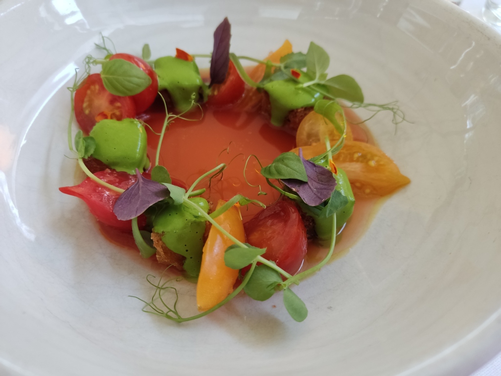 A bowl of bright red liquid circled by slices of red and yellow tomatoes with micro herbs and bright green sauce