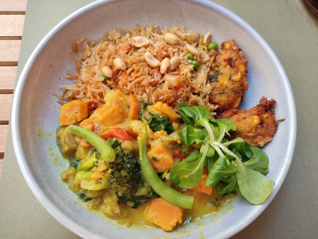 A bowl of rice with peanuts and peas, vegetable curry including green beans, broccoli and sweet potato, and two small sweetcorn fritters on the side
