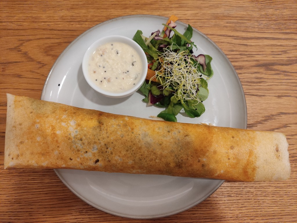 A plate holding a long dosa, a pot of coconut chutney, and a small fresh salad