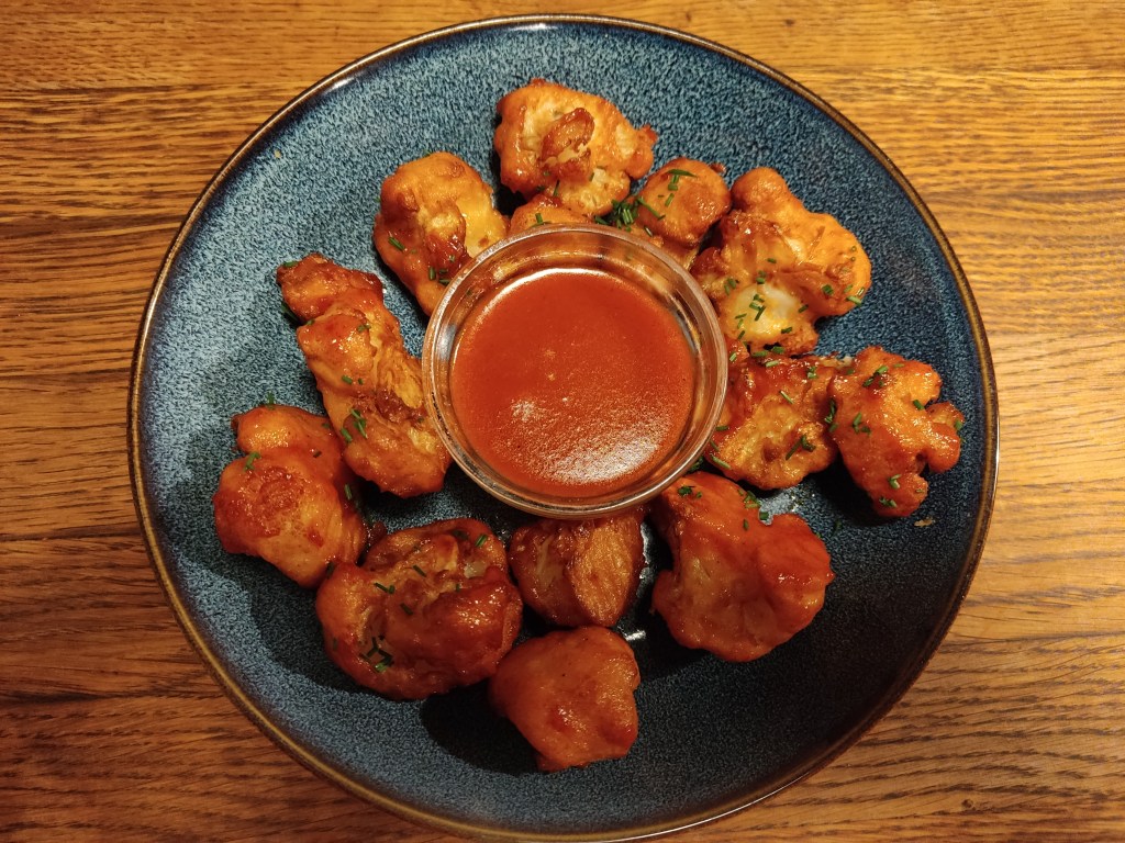 A plate with battered cauliflower pieces arranged a small pot of red dipping sauce