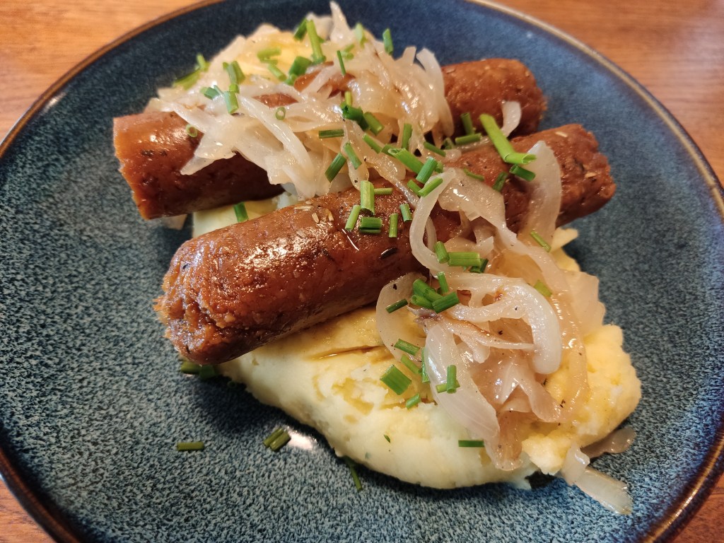 Two hearty sausages topped with soft fried onions sitting on top of creamy mashed potato