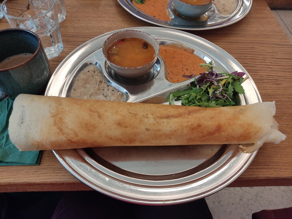 A metal try holding a long dosa, coconut chutney, a small soup, a sauce, and some fresh salad