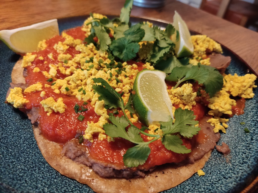 Three small tortillas topped with refried beans, tomato sauce, tofu scramble, lime wedges, and fresh coriander