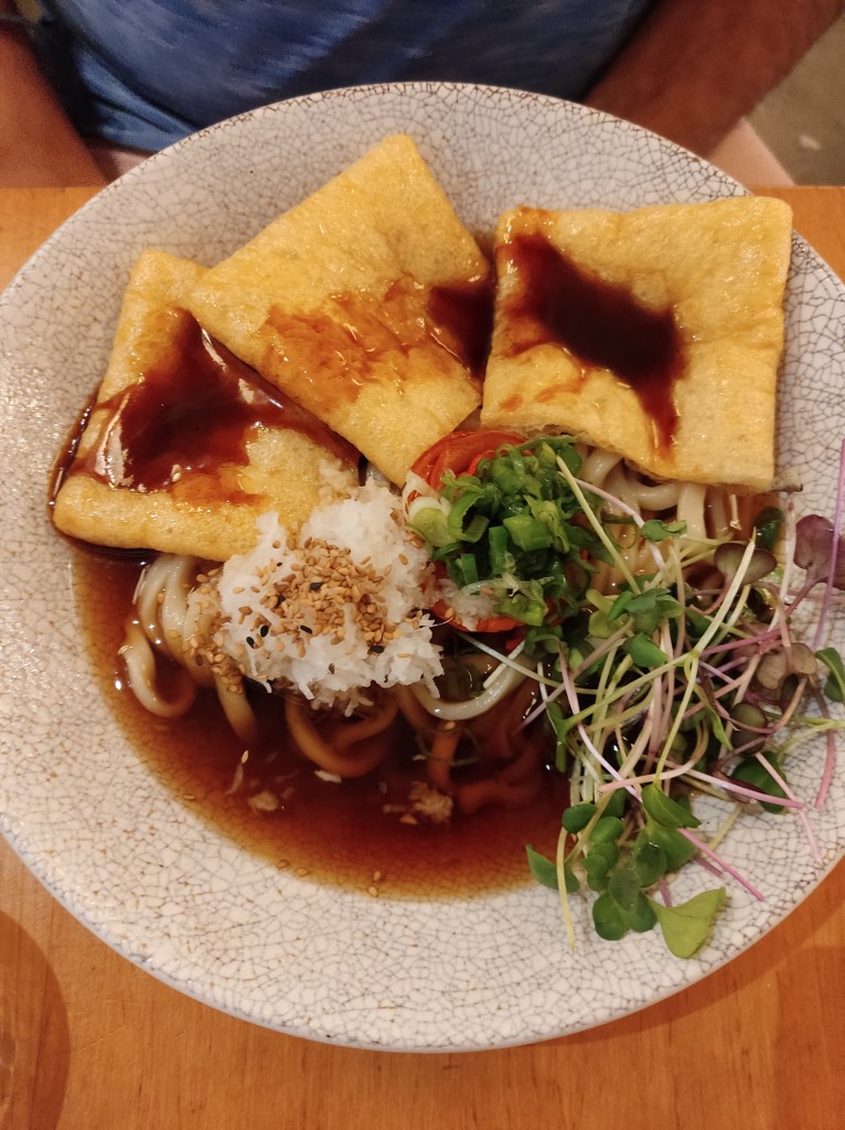 A bowl or clear dark brown broth with thick udon noodles, three big slices of fried tofu covered in sauce, grated radish, tomtato, spring onions, and greenery