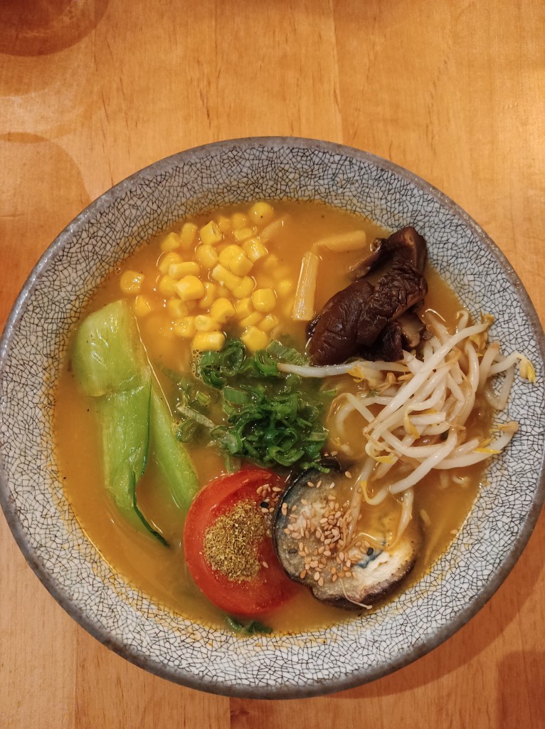 A bowl of ramen with a creamy yellowish broth topped with bok choy, sweetcorn, mushroom, aubergine, tomato, beansprouts, and spring onion