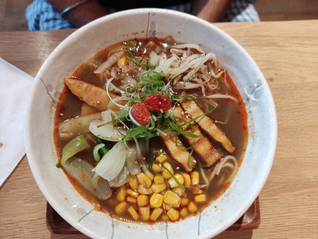 A bowl of brown broth filled with thin noodles, sweetcorn, tofu, bamboo, beansprouts, and other garnishes