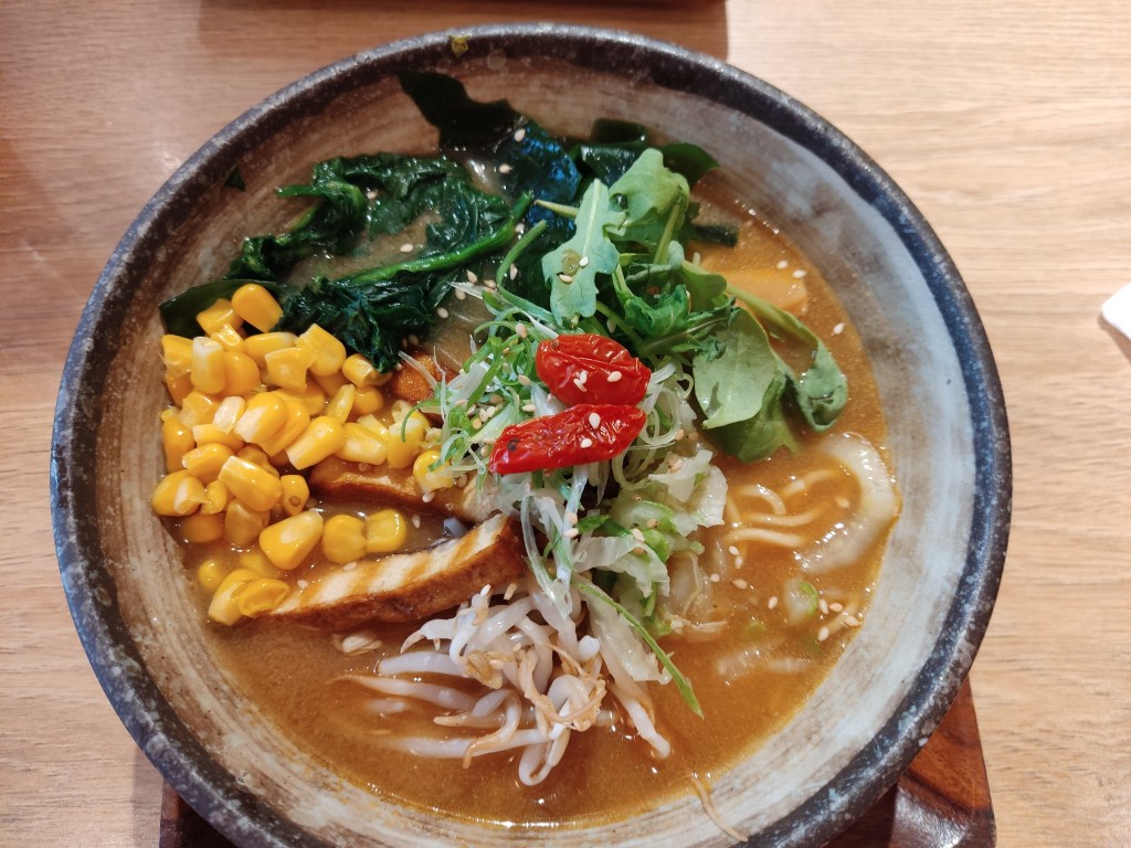 A bowl of light brown broth filled with thin noodles, sweetcorn, tofu, beansprouts, and other garnishes