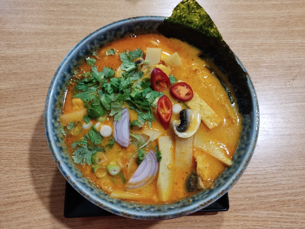 A bowl of vivid yellow broth garnished with seaweed, bamboo, mushroom, red onion, chilli, and lots of coriander