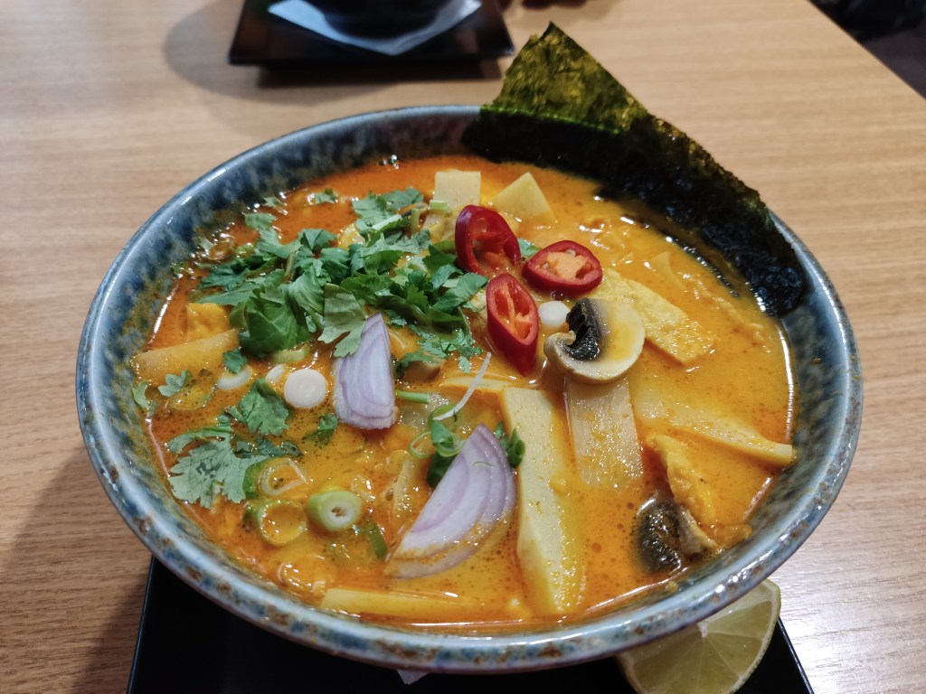 A bowl of vivid yellow broth garnished with seaweed, bamboo, mushroom, red onion, chilli, and lots of coriander