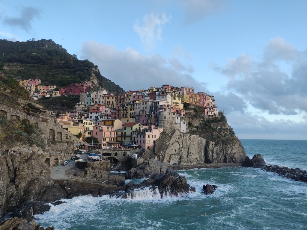 The small town of Manarola, lots of colourful buildings and a green hill rising up from a frothy blue sea