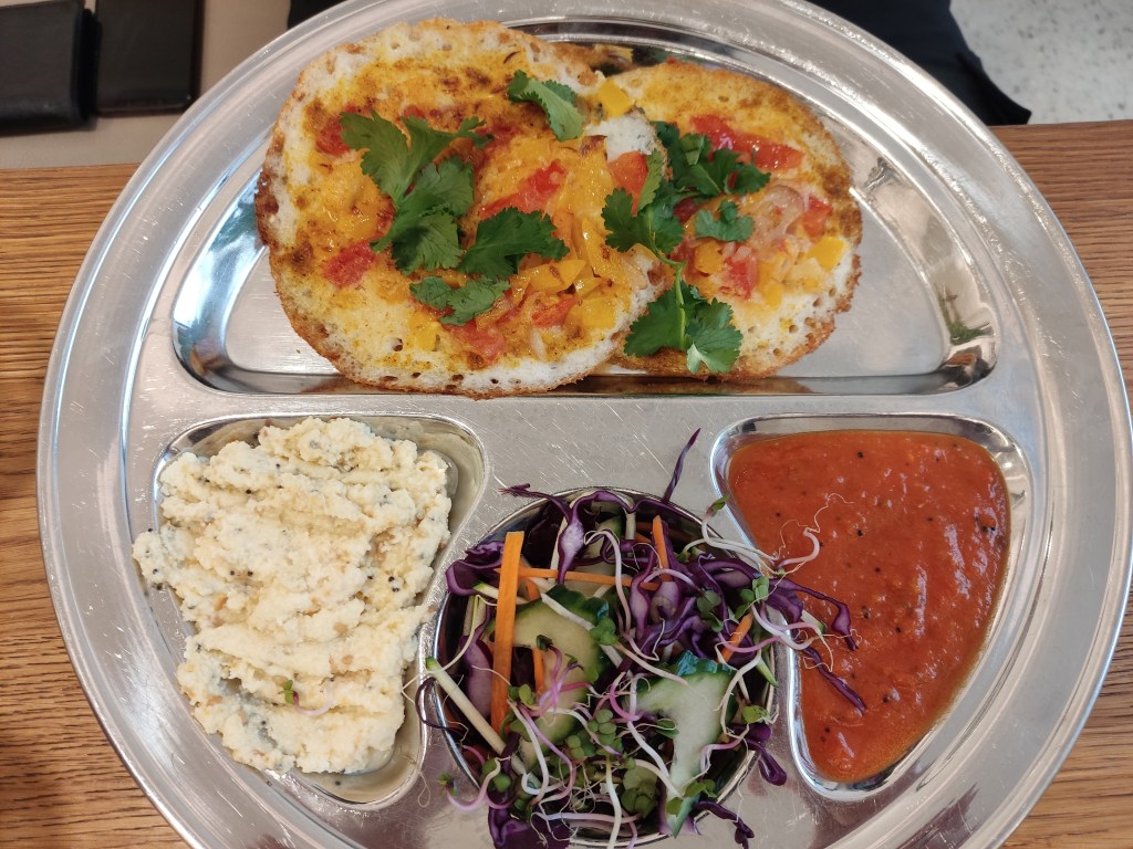 A metal tray with two small round pancakes topped with peppers and fresh coriander, and sections filled with coconut chutney, fresh salad, and a red sauce
