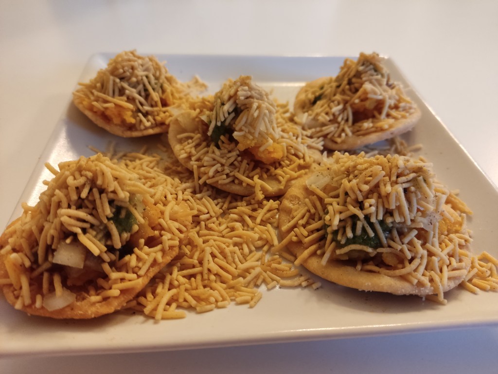 Five pieces of sev puri on a plate - a kind of cracker topped with little mounds of chutney, all buried under crispy sev