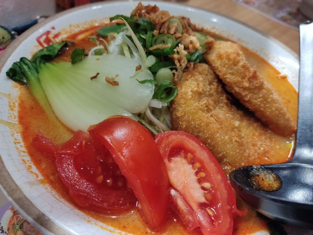 A bowl of almost orange broth topped with bok choy, tomato wedges, sugar snap peas, two fried croquettes, and various other garnishes