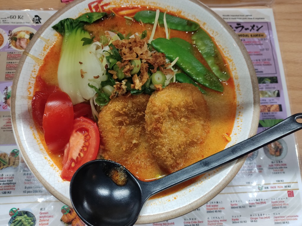 A bowl of almost orange broth topped with bok choy, tomato wedges, sugar snap peas, two fried croquettes, and various other garnishes