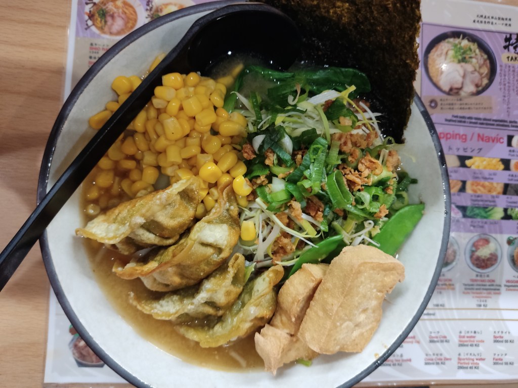 A bowl of beige broth topped with four crispy fried gyoza, two big chunks of fried tofu, lots of sweetcorn, and various other garnishes