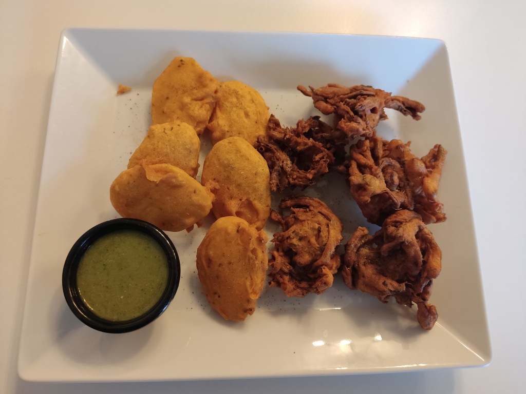 A large plate containing five onion pakora, dark brown in colour and wild shapes, and five potato pakora, uniform round shapes in golden batter. There's also a pot of green dipping sauce.