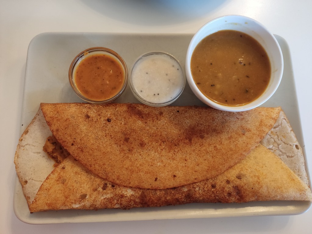 A crispy looking dosa with a small pot of soup and two dipping sauces