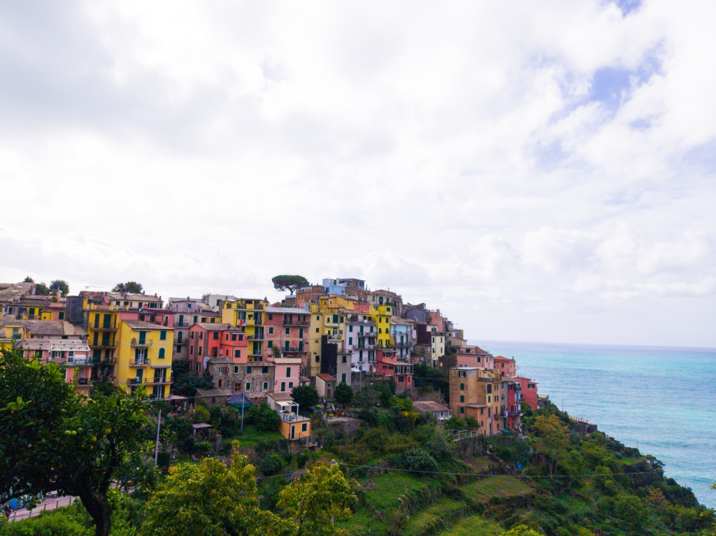 The small town of Corniglia, colourful buildings on a grassy hill with the sea behind it and a cloudy white sky overhead