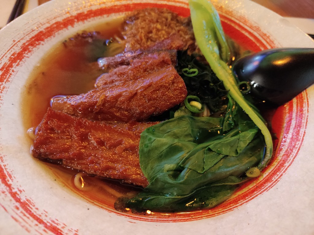 A bowl filled with clear brown broth and thin noodles, topped with bok choy and lots of slices of vegan bacon