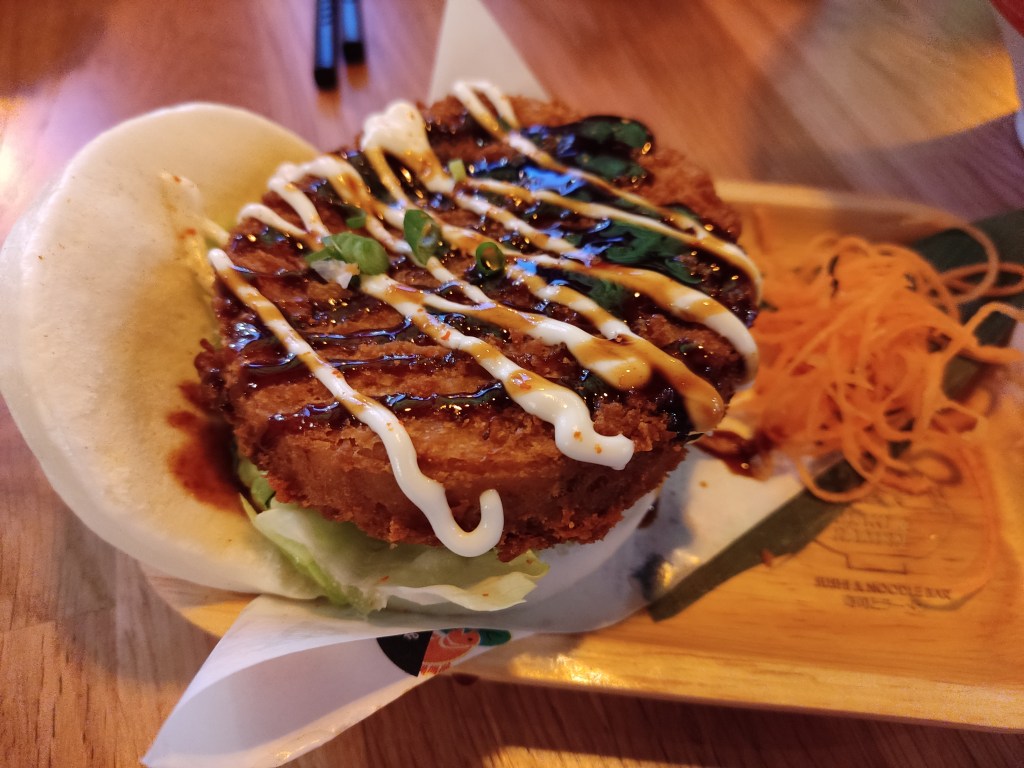 An open bao bun with a fried vegan chicken patty inside, drizzled with red and white sauces, along with some garnish.