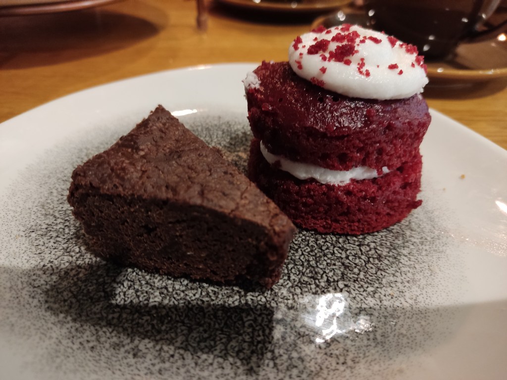 A triangular slice of brownie and an individual red velvet cake which is sandwiched and topped with a white cream.