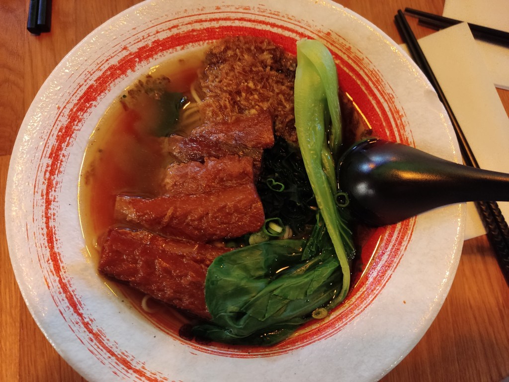 A bowl filled with clear brown broth and thin noodles, topped with bok choy and lots of slices of vegan bacon