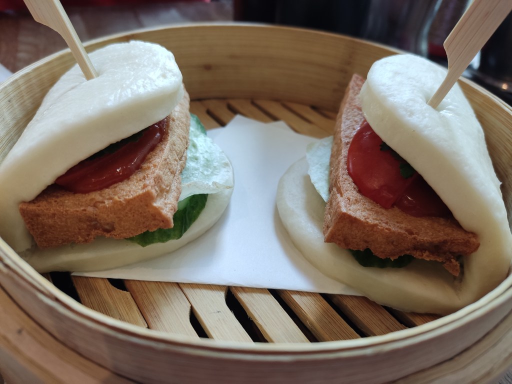Two bao buns sitting in a steamer, each containing a big slab of tofu with some lettuce and cucumber visible underneath and red sauce on top.