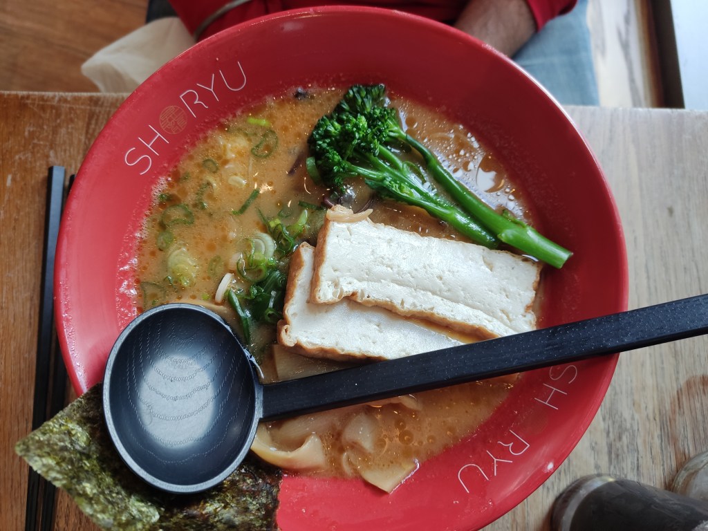 A big bowl of creamy light brown broth topped with seaweed, large slabs of tofu, tenderstem broccoli, and bamboo.