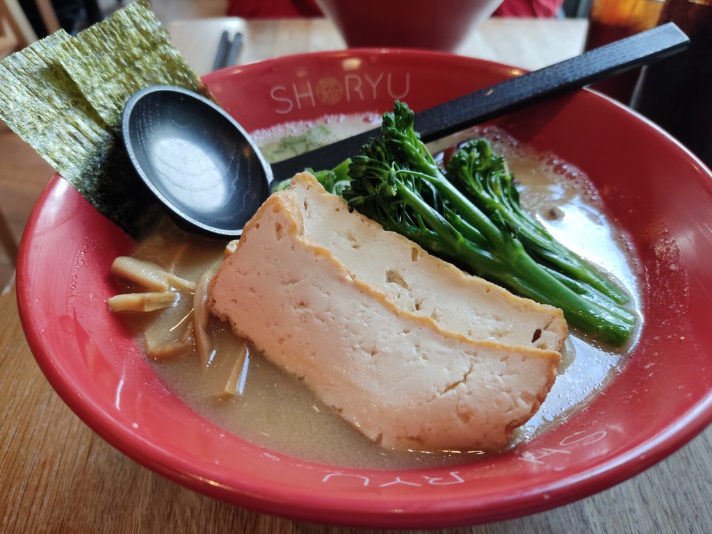 A big bowl of creamy broth topped with seaweed, large slabs of tofu, tenderstem broccoli, and bamboo.