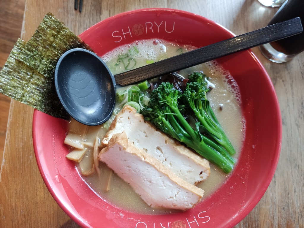 A big bowl of creamy broth topped with seaweed, large slabs of tofu, tenderstem broccoli, and bamboo.