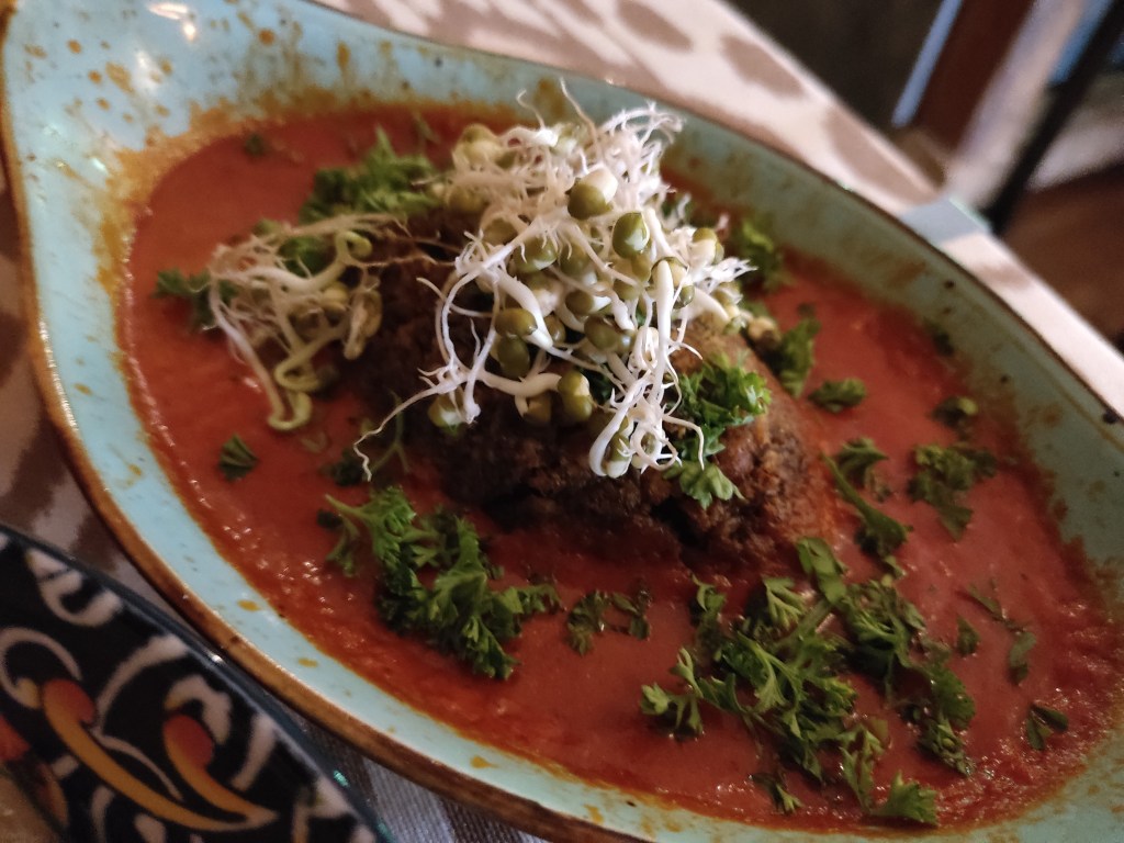 A large kofta sitting in a red sauce, garnished with bean sprouts and fresh herbs