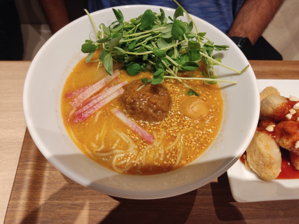 A bowl of ramen with a thick golden broth, a big meaty ball in the middle, lots of microgreens around, a blob of tahini, and loads of sesame seeds