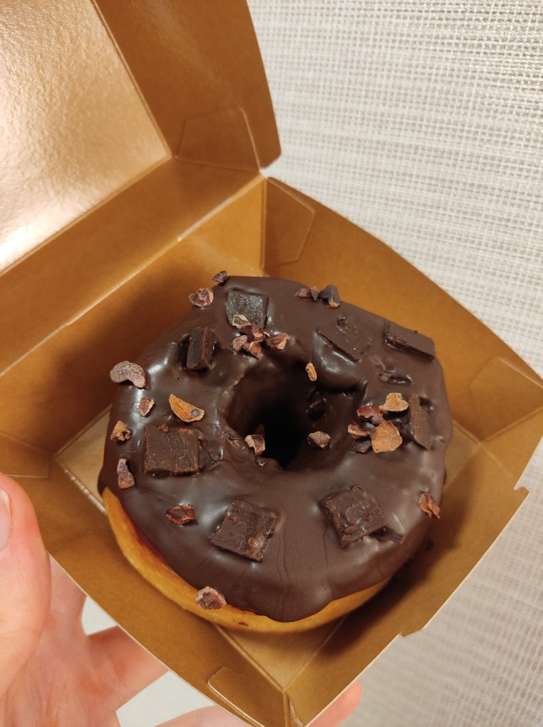 A doughnut with a thick chocolate coating and topped with some chocolate chunks and shavings, in a cardboard takeaway box