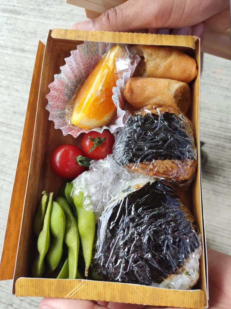 A disposable box containing two rice balls, some edamame, cherry tomatoes, a spring roll sliced in half, and an orange wedge