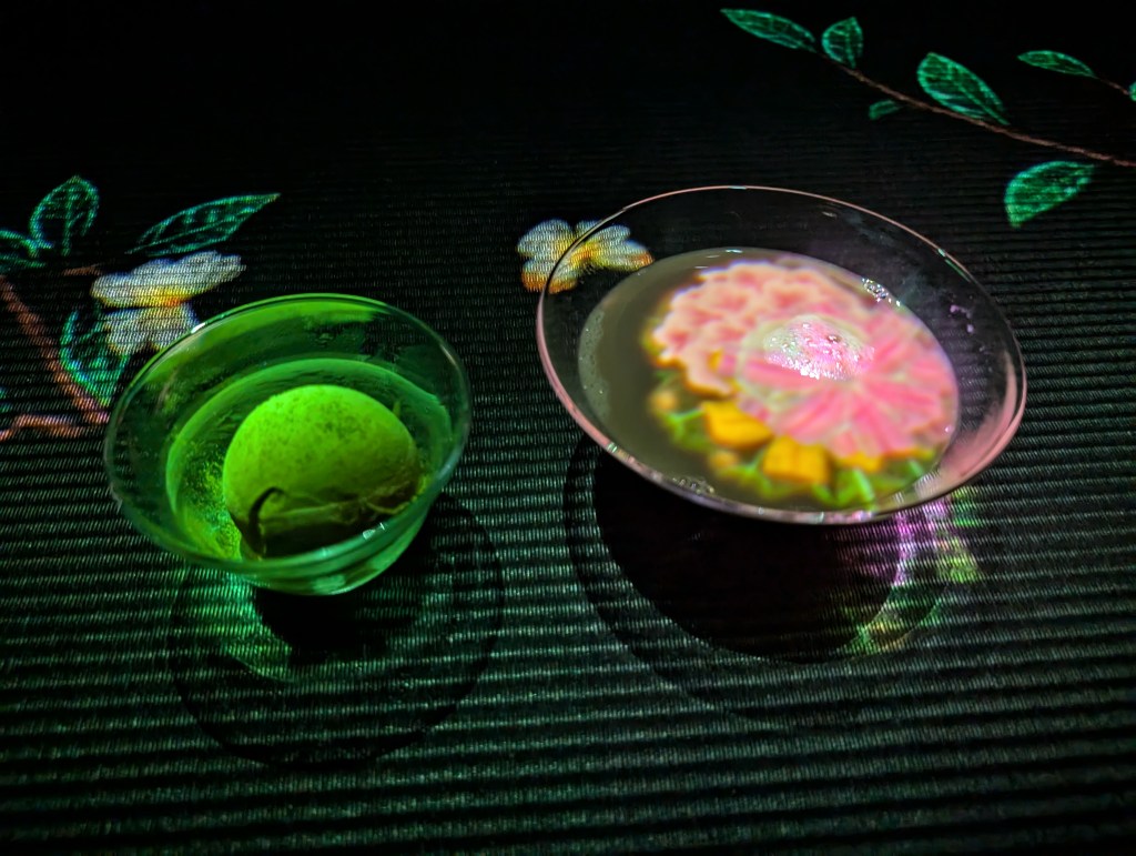 A bowl containing a scoop of green ice cream and a wide glass bowl containing tea. They are on a black table, and there are flowers, leaves, and butterflies projected on and around the items.