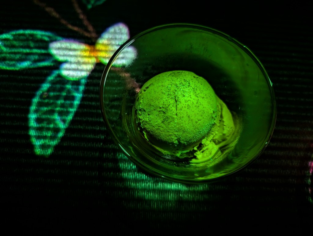 A bowl containing a scoop of green ice cream is placed on a black table, and there are leaves and butterflies projected on and around the it.