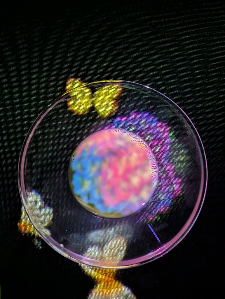 A wide glass bowl containing tea placed on a black table, and there are flowers and butterflies projected on and around the glass.