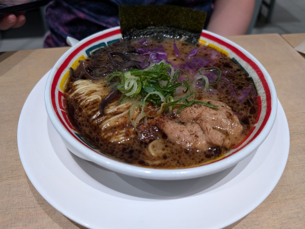 A bowl of ramen with a dark brown broth, a big portion of thin noodles, a few pieces of mock meats, loads of red cabbage and greens on top.
