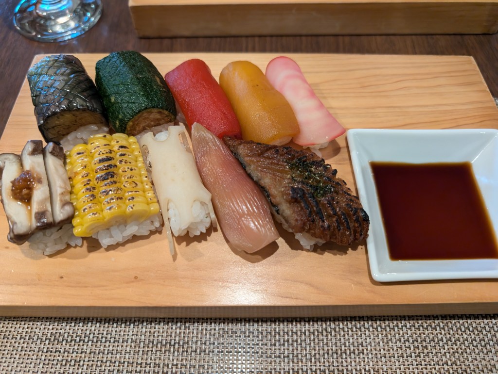 A board with ten pieces of sushi, mostly topped with with different vegetables, and a small dish of soy sauce