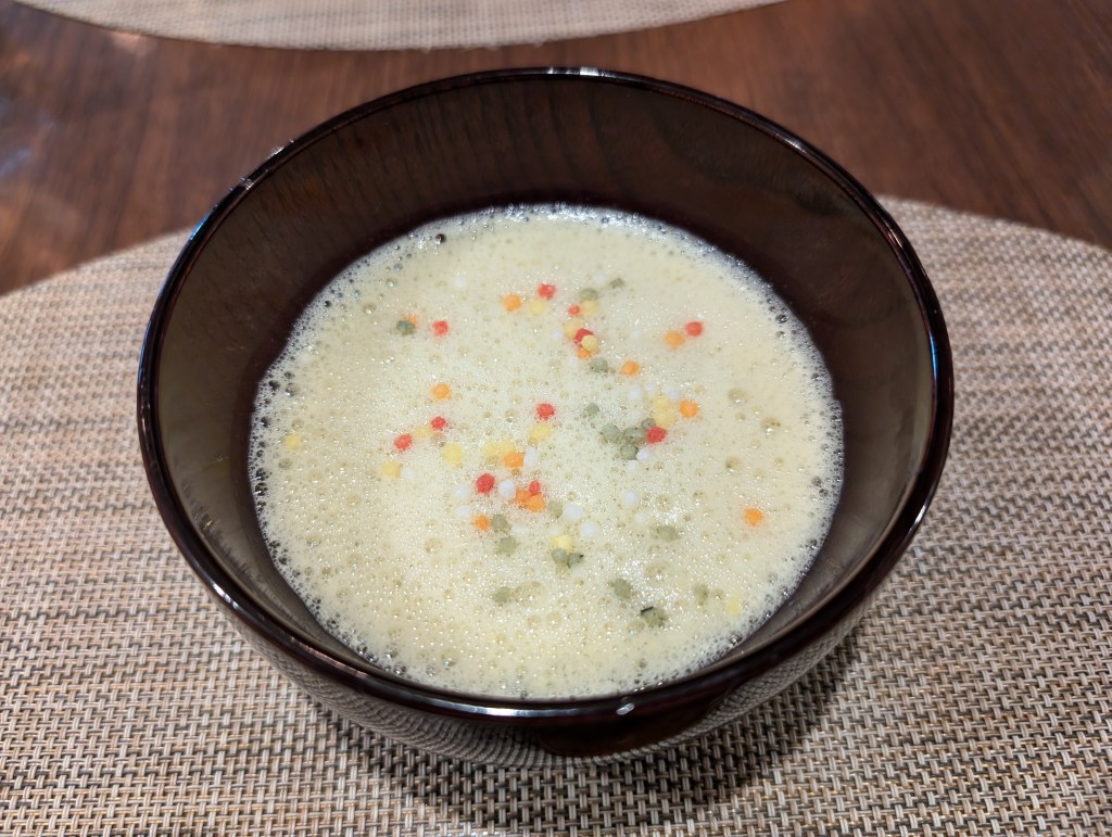 A small black bowl filled with a frothy, creamy, pale yellow soup. There are pieces of orange, green, and red puffed rice.