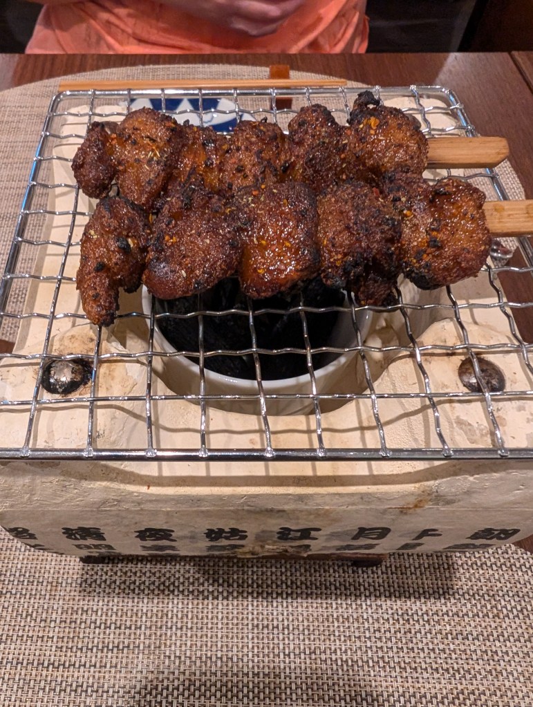 Two skewers of vegan meat, nicely charred and sticky looking. They are on a metal grill over a heating device at the table.