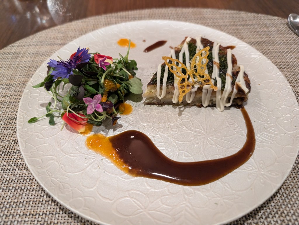 A plate with a slice of okonomiyaki, drizzled with mayo and dressed with sauce. There's a colourful and floral salad on one side. 