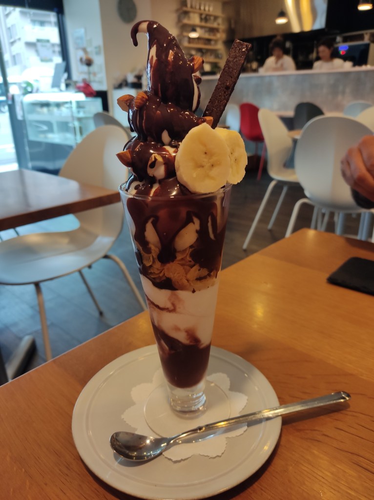 A tall glass filled with layers of ice cream, chocolate sauce, banana, and cornflakes. The top is adorned with chocolate sauce, almond, banana slices, and a chocolate finger